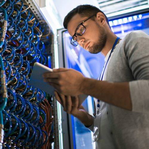 Low angle  portrait of young man using digital tablet standing by server cabinet while working with supercomputer in blue light